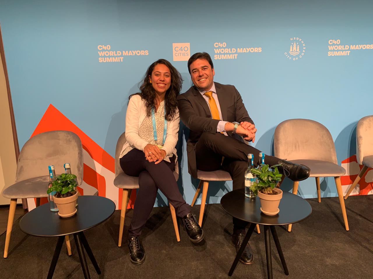 Secretário Luiz Alvaro e Coordenadora Marina Andrade, ambos sentados em cadeiras e de pernas cruzadas. Ao fundo, um painel azul com o texto "C40 World Mayors Summit" em branco.